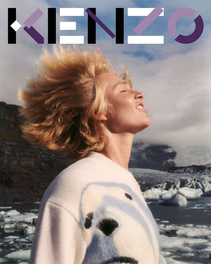 Fashion portrait with adult woman in patterned sweater, blonde hair blowing, mountain and lake backdrop, featuring Kenzo logo.