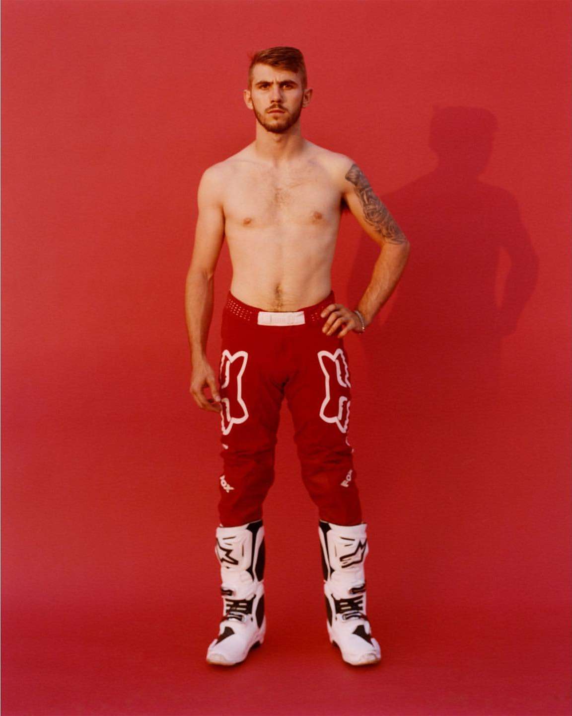 Full-length portrait of a shirtless man in red motocross pants and white boots standing against a solid red background in a photo studio.