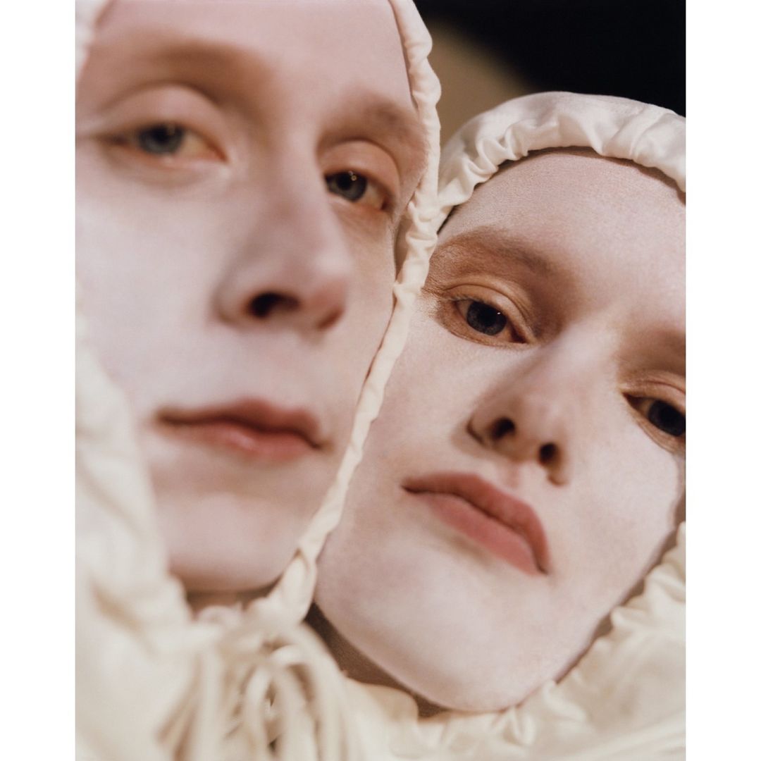 Close-up of two individuals with cream-colored face makeup and head coverings, faces close together, in a softly lit, intimate studio portrait.