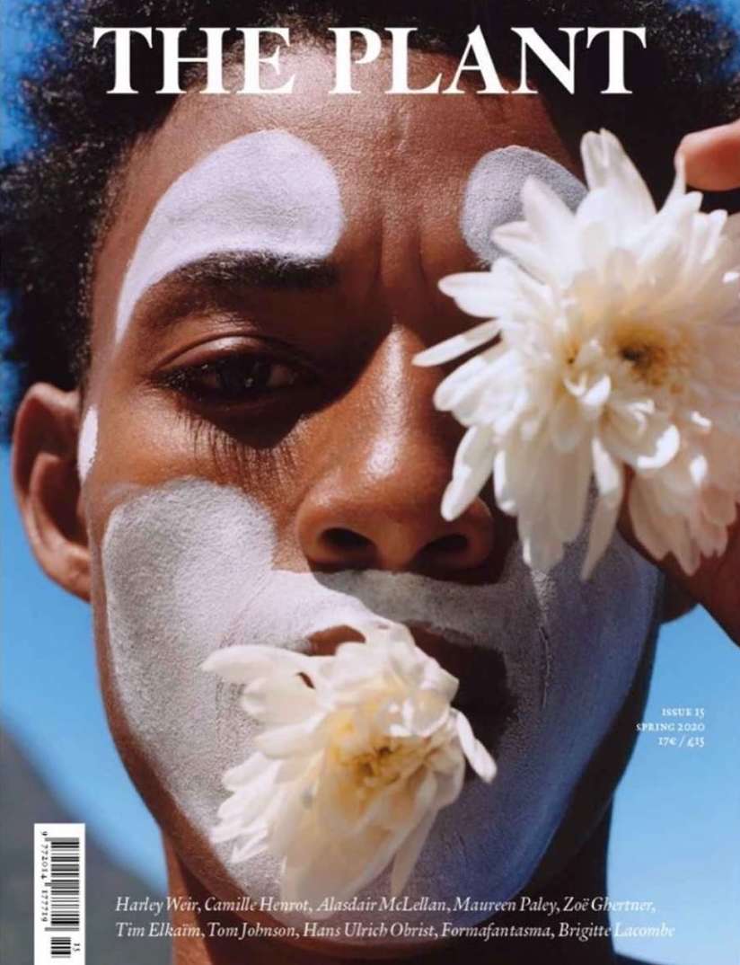 Magazine cover featuring a close-up portrait of a dark-skinned male model with white floral face paint and holding white flowers.