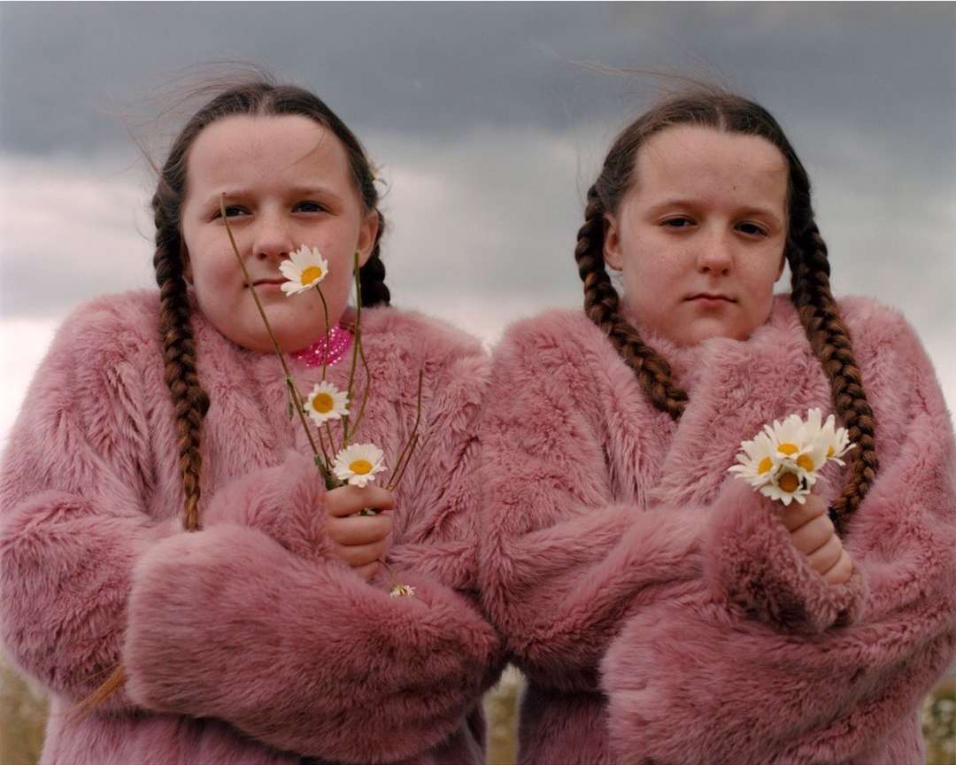 Twin girls in pink fur coats stand outdoors, holding daisies against a cloudy sky, creating a whimsical and slightly surreal portrait.