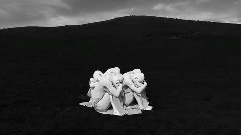 Monochromatic outdoor art photography features a huddled group of people in a field with a dark hill in the background under a cloudy sky.