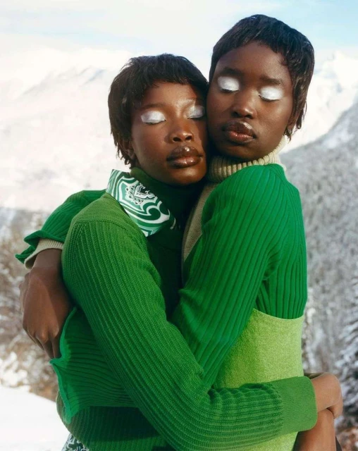 Two dark-skinned women embrace in green outfits against a snowy mountain backdrop, their eyes are covered with white makeup.
