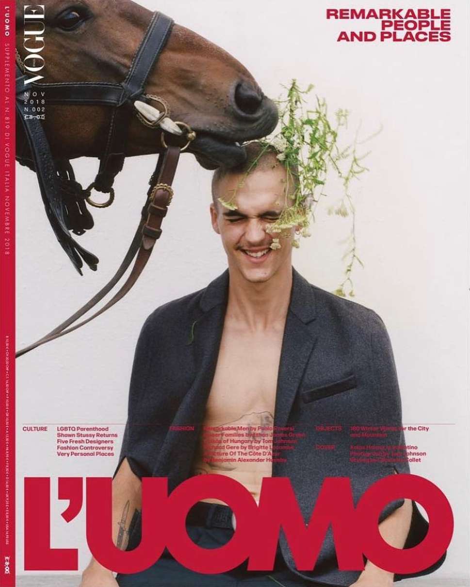 Man smiling with flowers tucked in his hair and a horse nuzzling his head against a white background