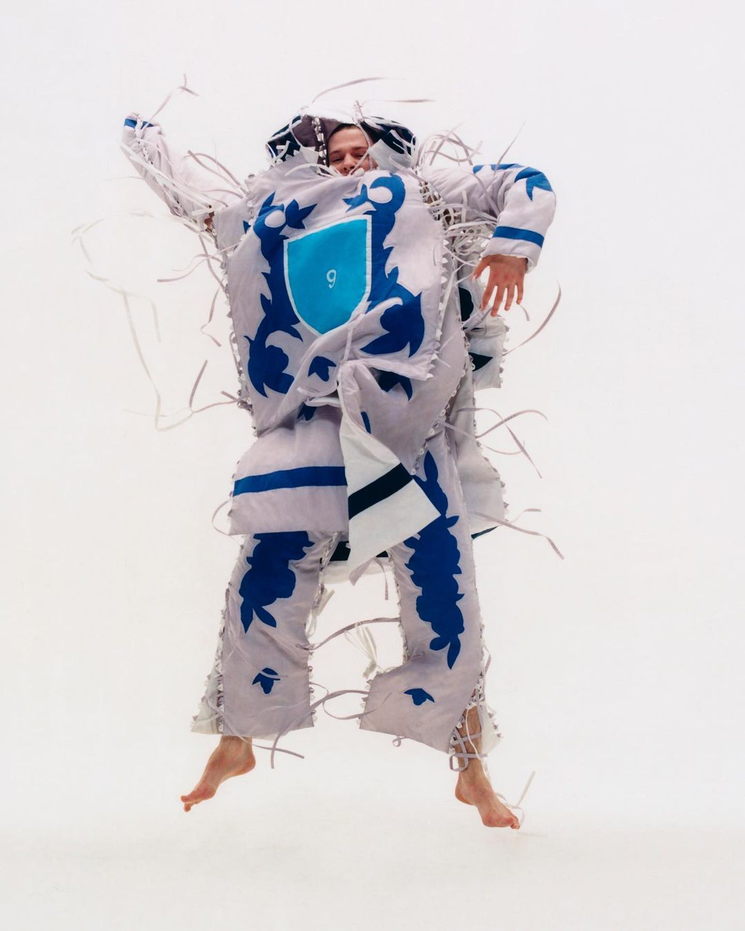 Person jumping in a white and blue ornate costume with flowing white streamers against a bright white backdrop in a studio setting.