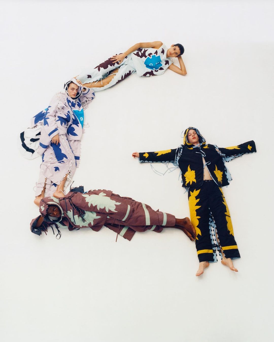 Four models in patterned outfits lying on a white background, arranged to suggest a letter, showcasing fashion and creative posing.