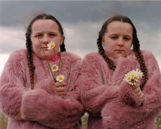 Two young girls with braided hair wearing pink faux fur coats holding daisies under an overcast sky, by photographer Tom Johnson.