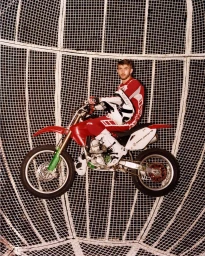 Motocross rider in red and white gear holding a helmet while seated on a red dirt bike inside a metal cage structure.