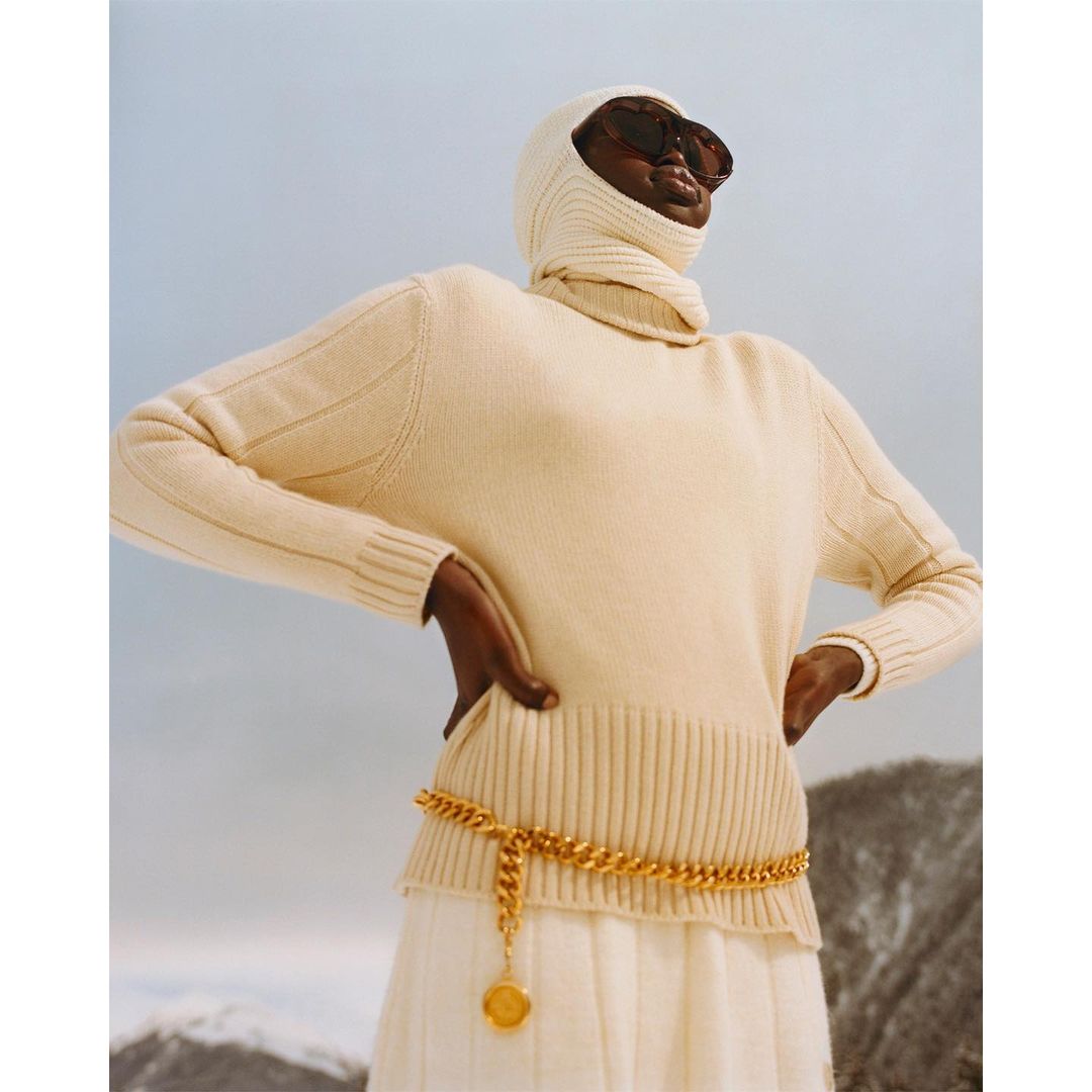 Model wearing a cream turtleneck sweater, matching balaclava, oversized sunglasses, and a gold chain belt outdoors against a mountain background.