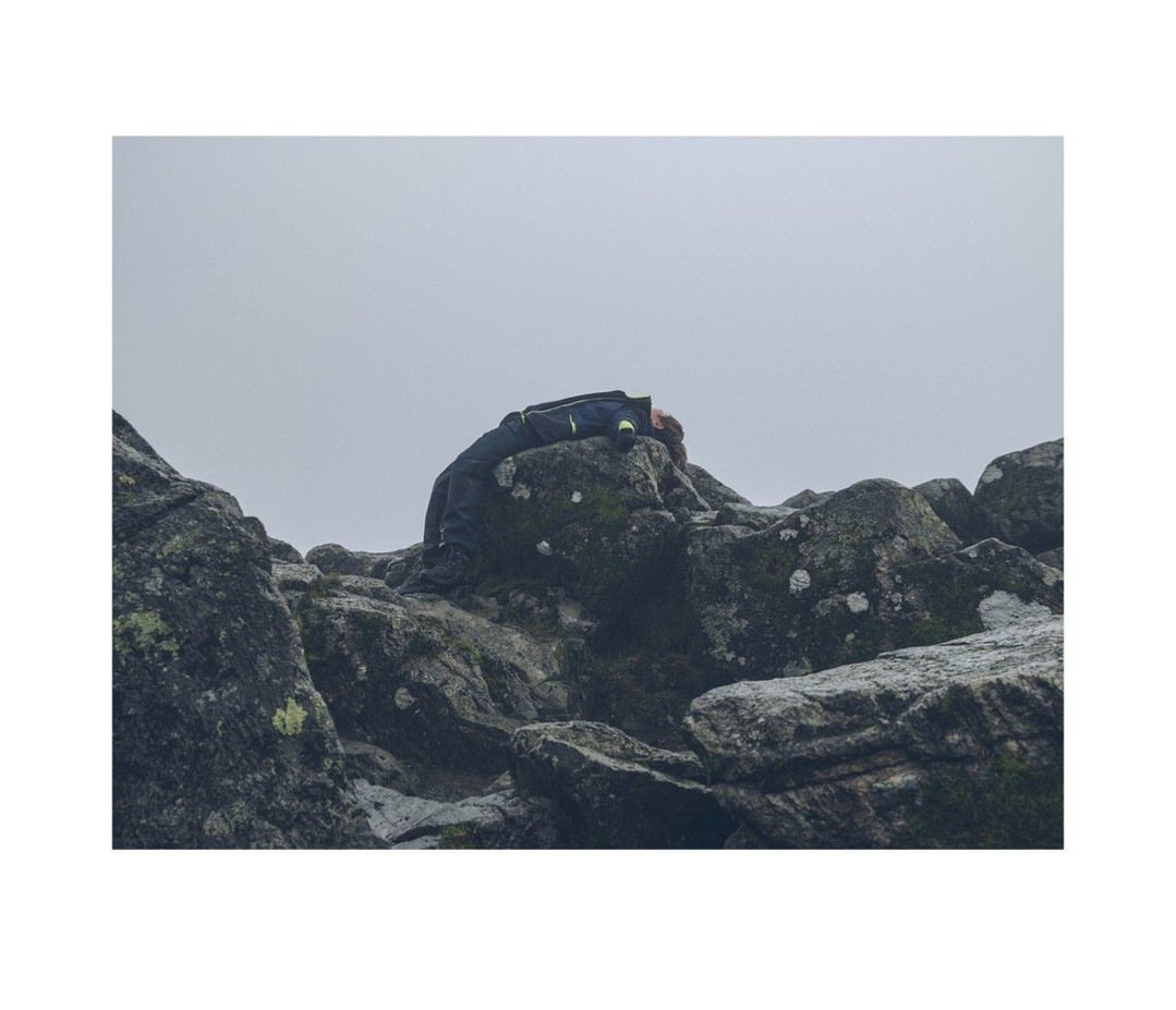 A person rests atop mossy rocks under an overcast sky, their dark jacket blending with the rugged landscape in a muted palette.