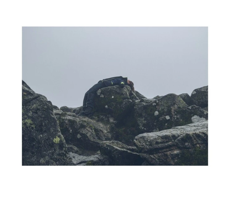 A person rests atop mossy rocks under an overcast sky, their dark jacket blending with the rugged landscape in a muted palette.
