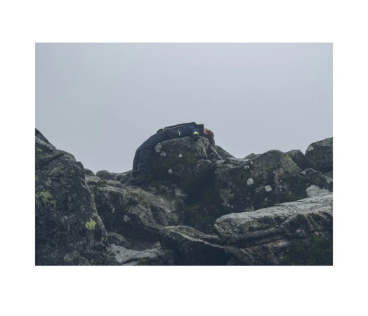 A person rests atop mossy rocks under an overcast sky, their dark jacket blending with the rugged landscape in a muted palette.