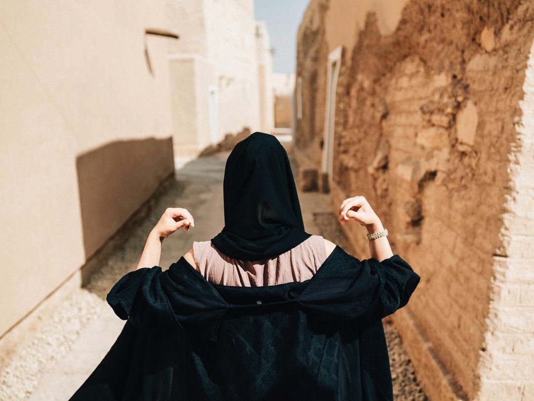 Woman with black headscarf walks through sandstone alleyway. Sunlight brightens the weathered building exterior in the background.