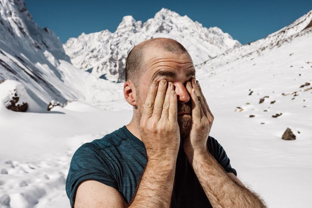 Bald adult male with beard rubs his face outdoors on a snowy hillside; mountain range visible in the background against a clear blue sky.