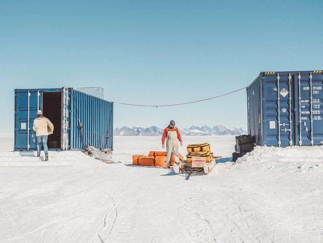 Two people stand near blue shipping containers in a snowy outdoor setting with distant mountains under a clear blue sky, possibly in Antarctica.