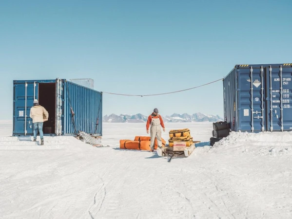 Two people stand near blue shipping containers in a snowy outdoor setting with distant mountains under a clear blue sky, possibly in Antarctica.