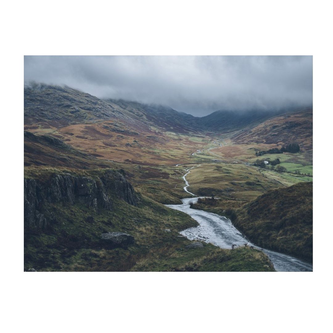 Scenic view of a river winding through a valley with mountains in the background under a cloudy sky, creating a peaceful landscape.