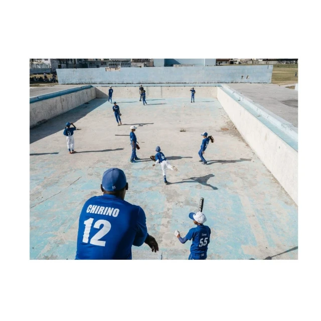 Youth baseball team practices outdoors on a concrete court. Players in blue and white uniforms throw and catch a ball.