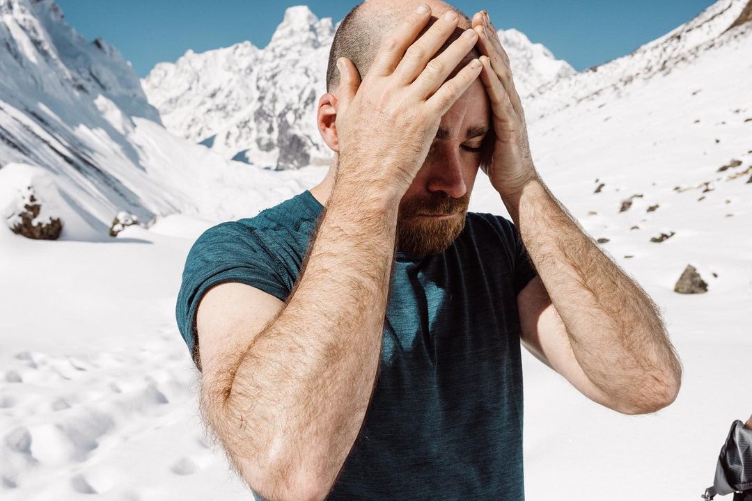 Bald man in blue shirt holds his head in a snowy mountain landscape with clear blue sky; outdoors, daytime, medium shot, natural light.