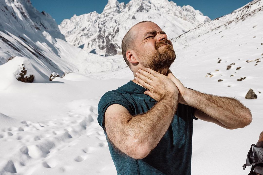 Adult male in a blue shirt touches his neck outdoors against a backdrop of snow-covered mountains under a blue sky in daylight.