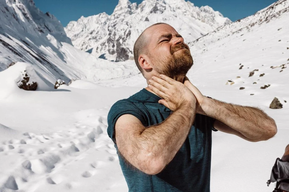 Adult male in a blue shirt touches his neck outdoors against a backdrop of snow-covered mountains under a blue sky in daylight.