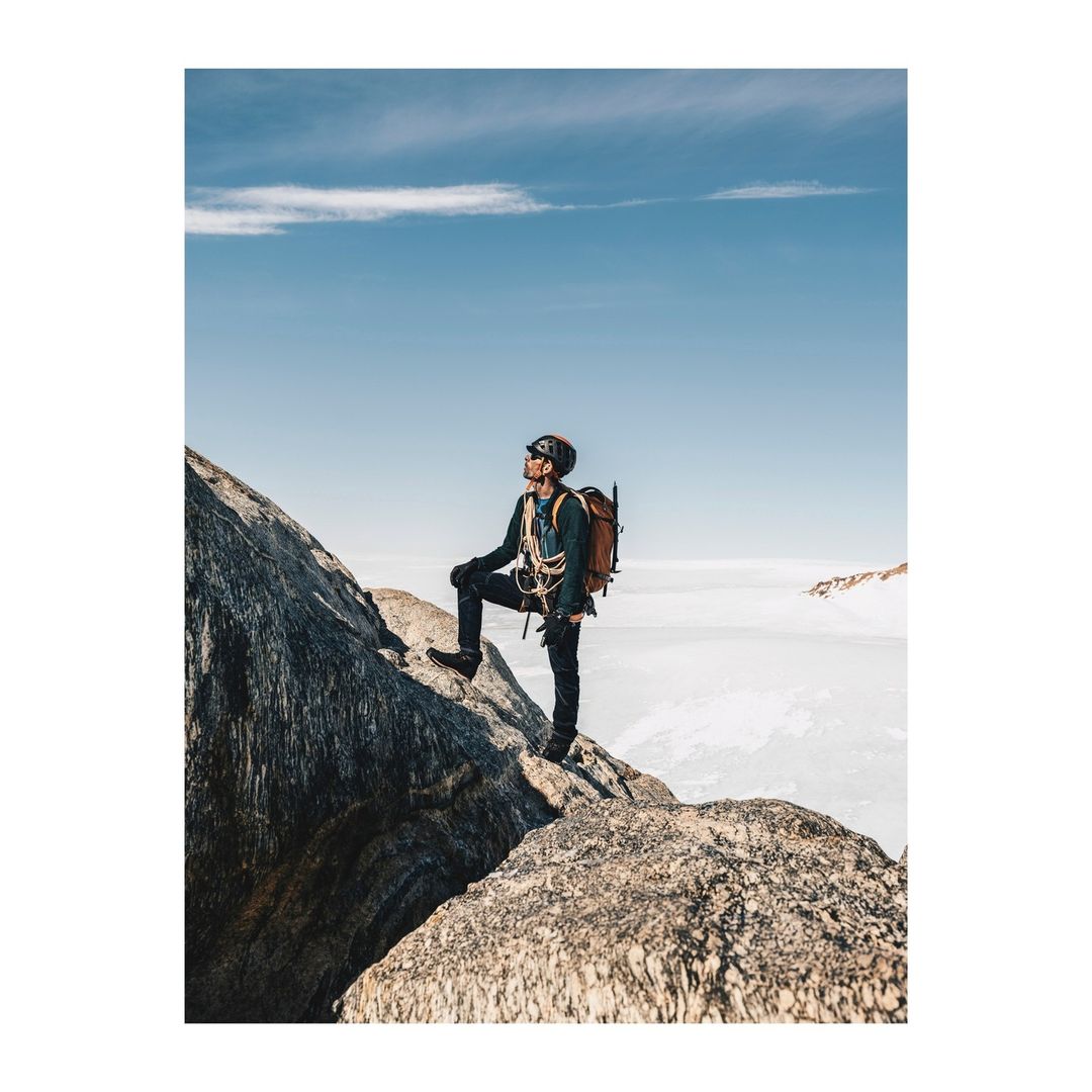 Man climbing mountain in outdoor adventure, wearing climbing gear, backpack and helmet, with a clear blue sky and cloudscape background.