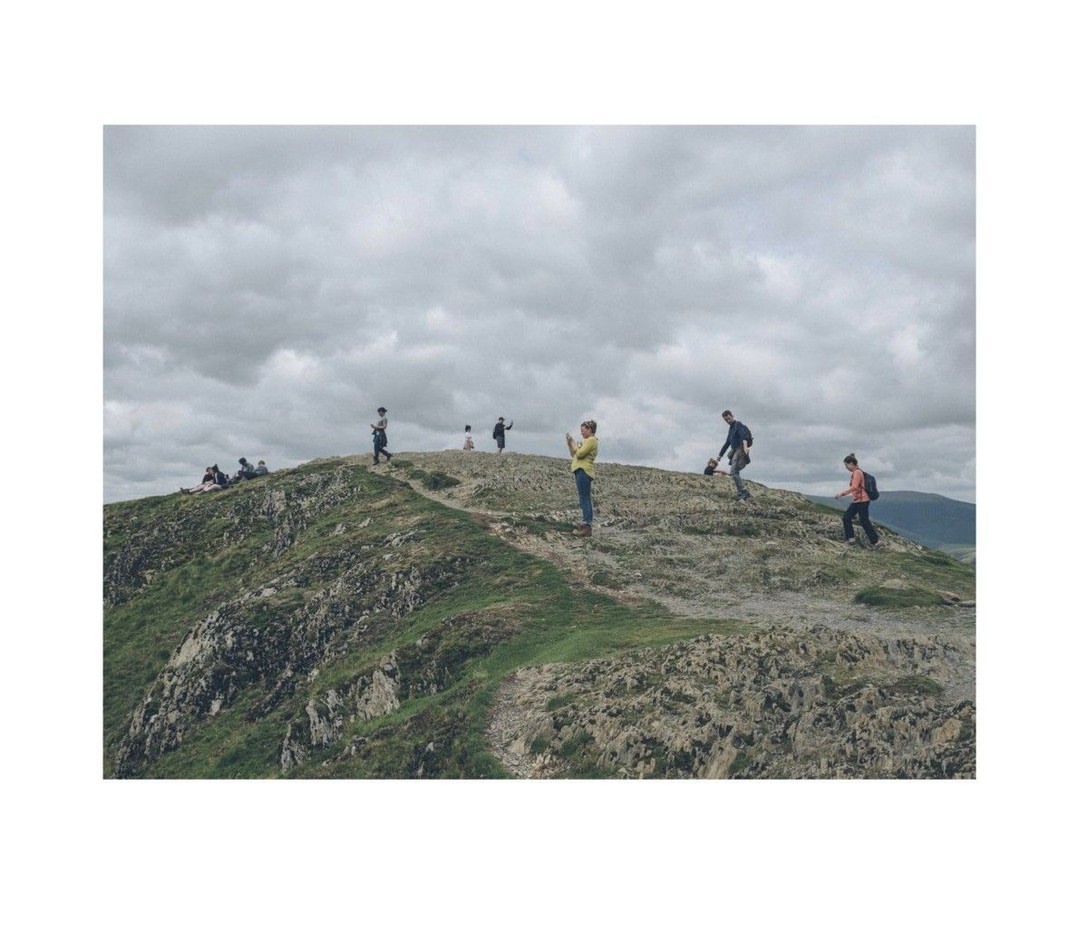 A group of people are scattered along the top of a grassy, rocky hill under a cloudy sky in a landscape orientation.