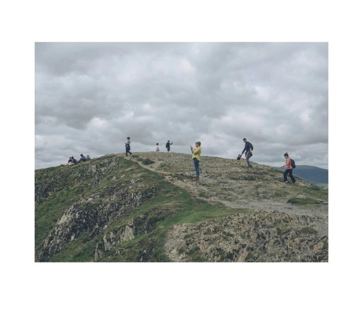 A group of people are scattered along the top of a grassy, rocky hill under a cloudy sky in a landscape orientation.
