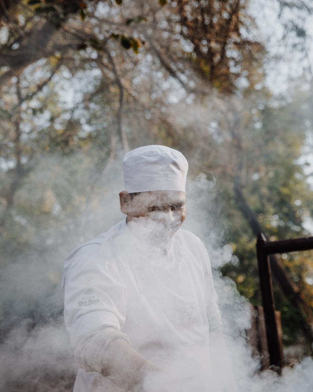 Chef in white uniform surrounded by smoke outdoors preparing food, with leafy trees visible in the background in a natural setting.
