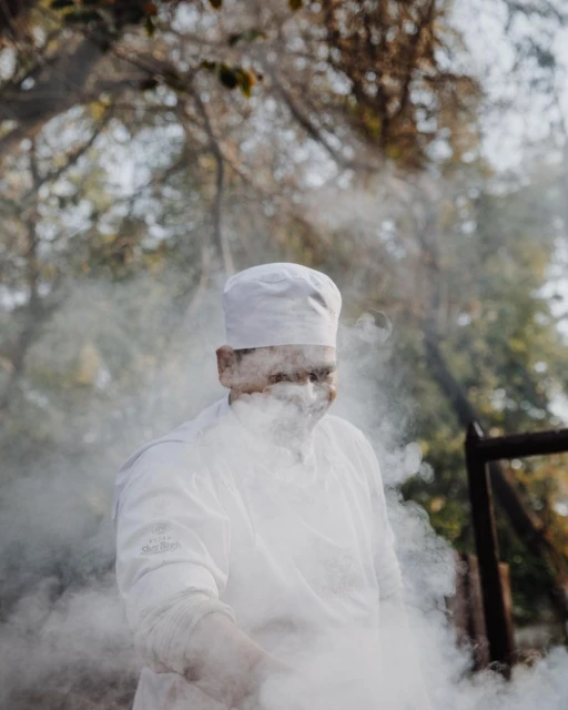 Chef in white uniform surrounded by smoke outdoors preparing food, with leafy trees visible in the background in a natural setting.