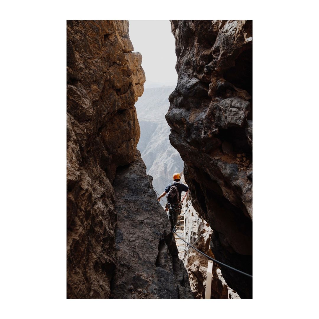 Man climbing via ferrata on rocky mountainside with canyon view, wearing an orange helmet, seen from behind in a medium shot.