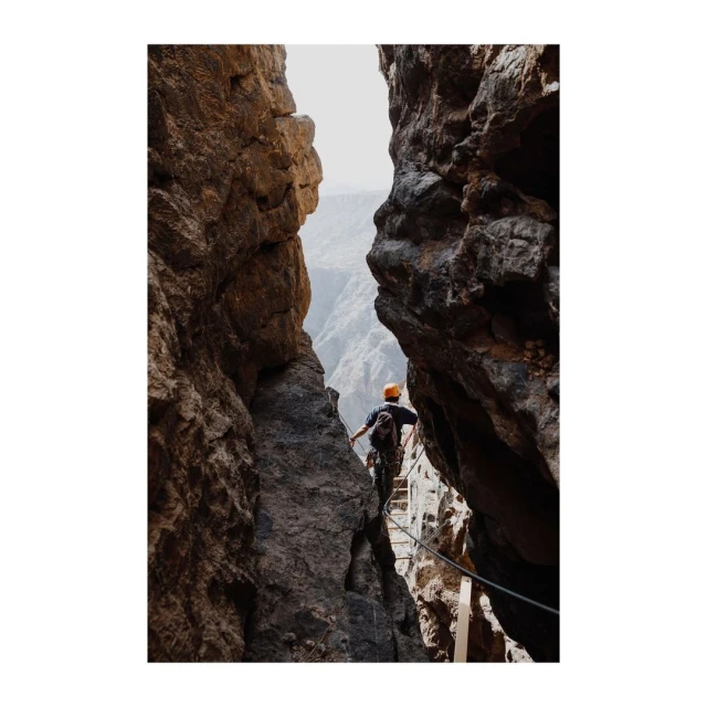 Man climbing via ferrata on rocky mountainside with canyon view, wearing an orange helmet, seen from behind in a medium shot.