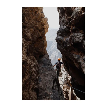 Man climbing via ferrata on rocky mountainside with canyon view, wearing an orange helmet, seen from behind in a medium shot.