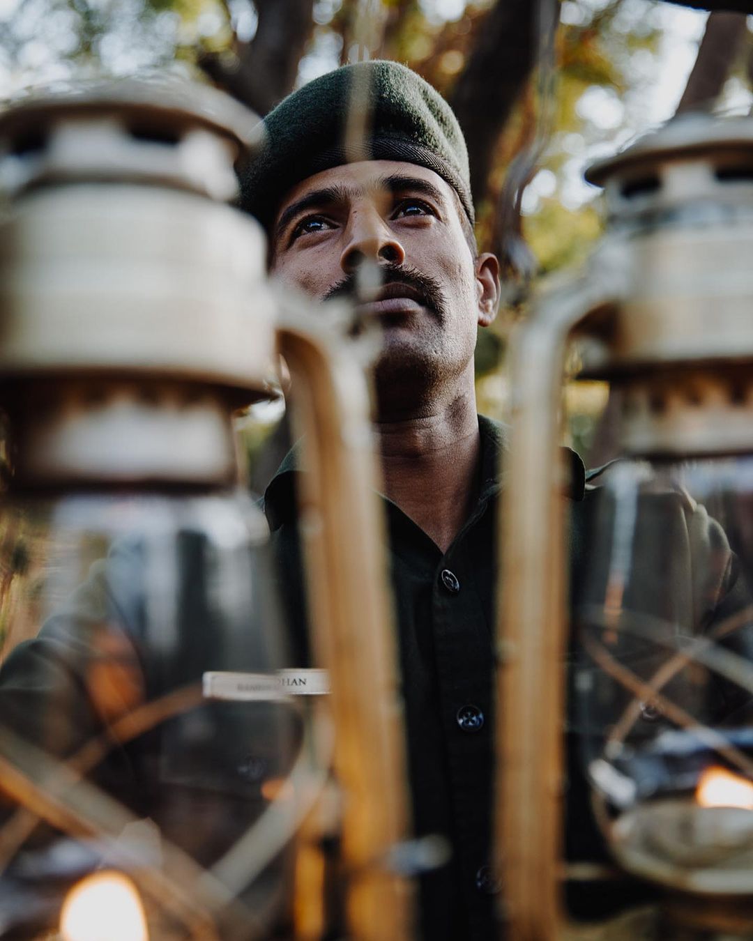 Portrait of a man in uniform looking upward, framed by lit kerosene lanterns, set against a blurred natural outdoor background.