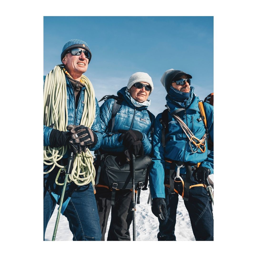 Three climbers stand on a snowy peak against a bright blue sky, wearing winter gear with ropes and walking sticks, enjoying a sunny day.