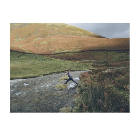 Person jumps over river rocks in rural valley with grassy hills and cloudy sky, captured in a medium shot with natural lighting.
