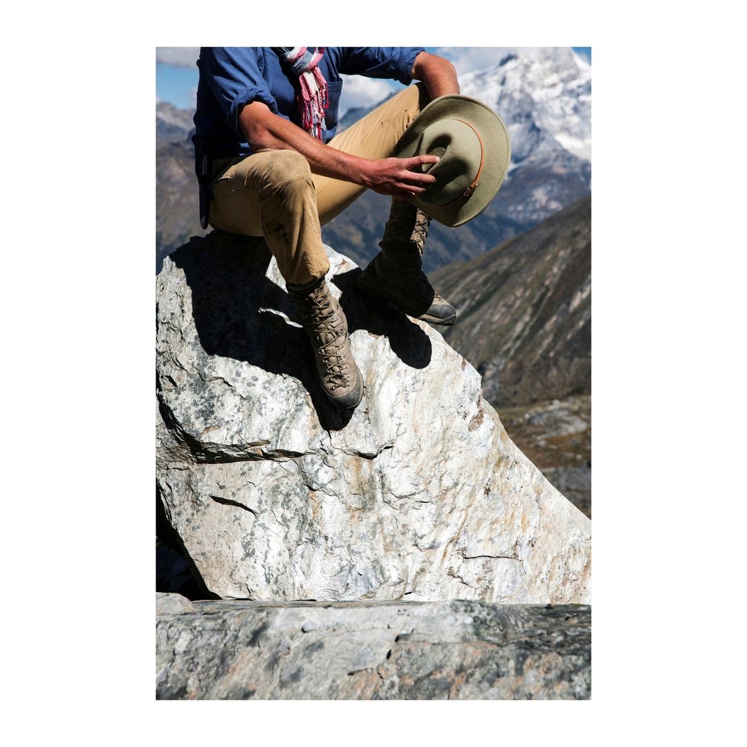 Man in outdoor boots and hat sits on a large rock formation in a mountain landscape, wearing casual clothing, enjoying the scenery.