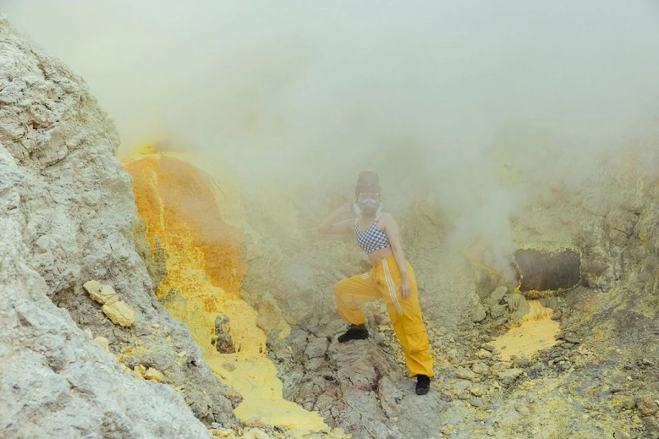 Woman with gas mask and hat poses on rocky terrain with yellow sulfur deposits surrounded by thick fog or smoke; wide, environmental shot.