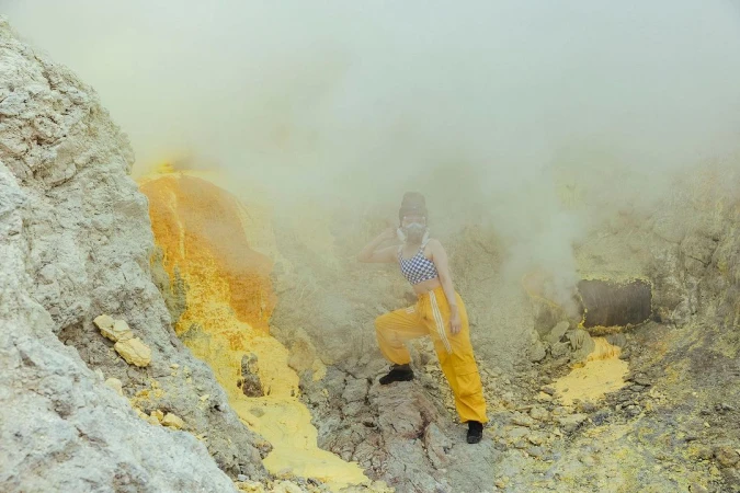Woman with gas mask and hat poses on rocky terrain with yellow sulfur deposits surrounded by thick fog or smoke; wide, environmental shot.