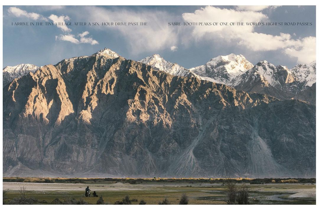 Majestic mountain range with snow-capped peaks under a partly cloudy sky, with a figure on a motorcycle in the foreground.