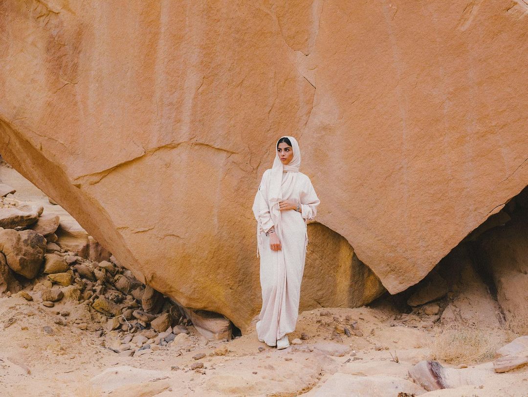 A woman in a light pink abaya and hijab stands in front of a large rock formation in the desert, creating a serene and natural portrait.