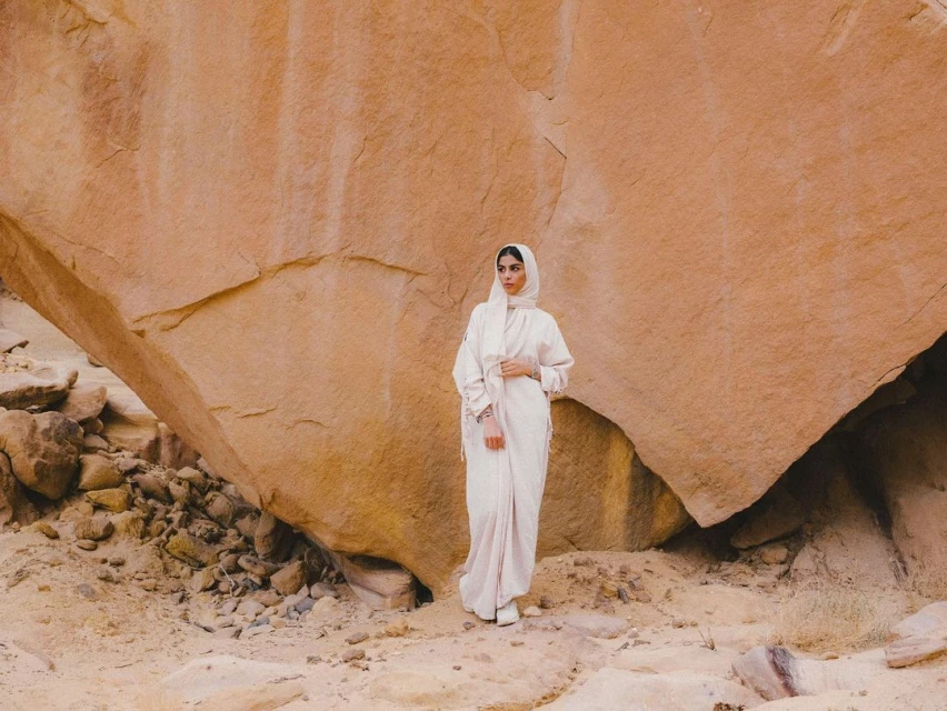 A woman in a light pink abaya and hijab stands in front of a large rock formation in the desert, creating a serene and natural portrait.