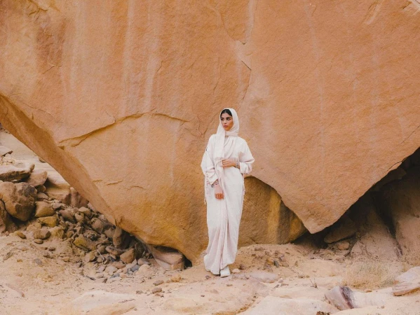 A woman in a light pink abaya and hijab stands in front of a large rock formation in the desert, creating a serene and natural portrait.