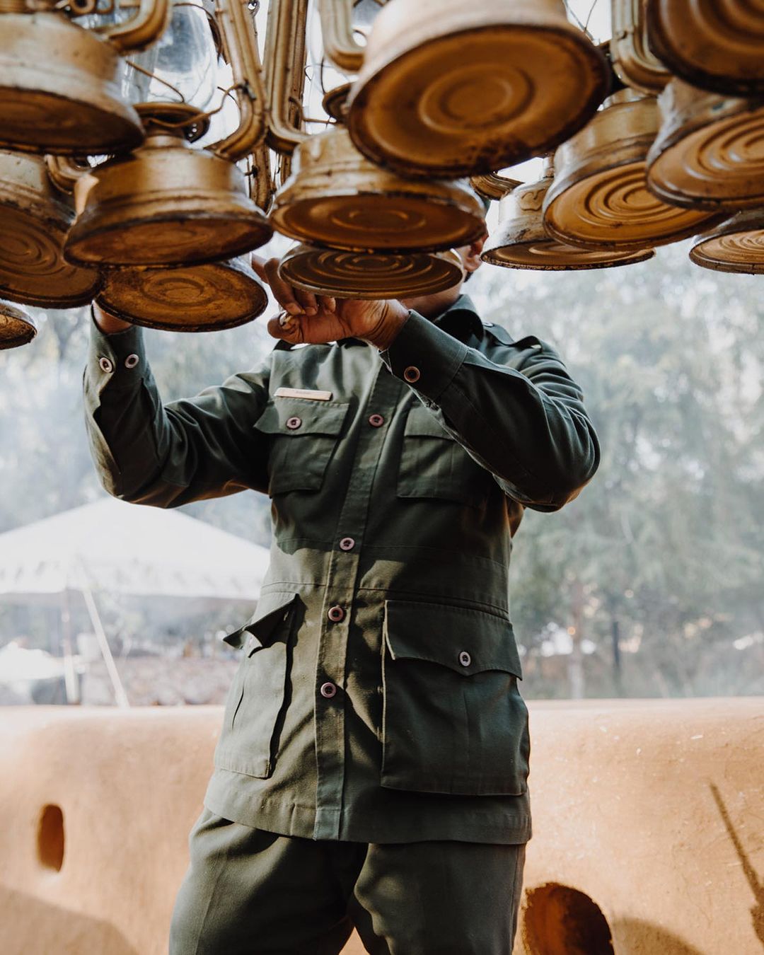 Person in green jacket adjusting antique brass chandelier outdoors, white tent in background, on a sunny day.