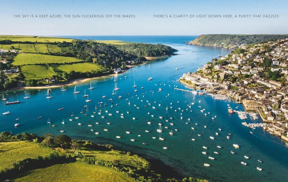 Aerial view of a coastal scene featuring a waterway with sailboats, surrounded by green hills and a village on the right side.
