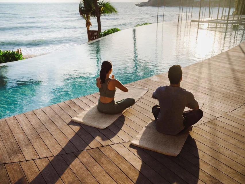 Woman and man practice yoga in lotus position on mats near an infinity pool overlooking the ocean at a tropical resort at sunrise.