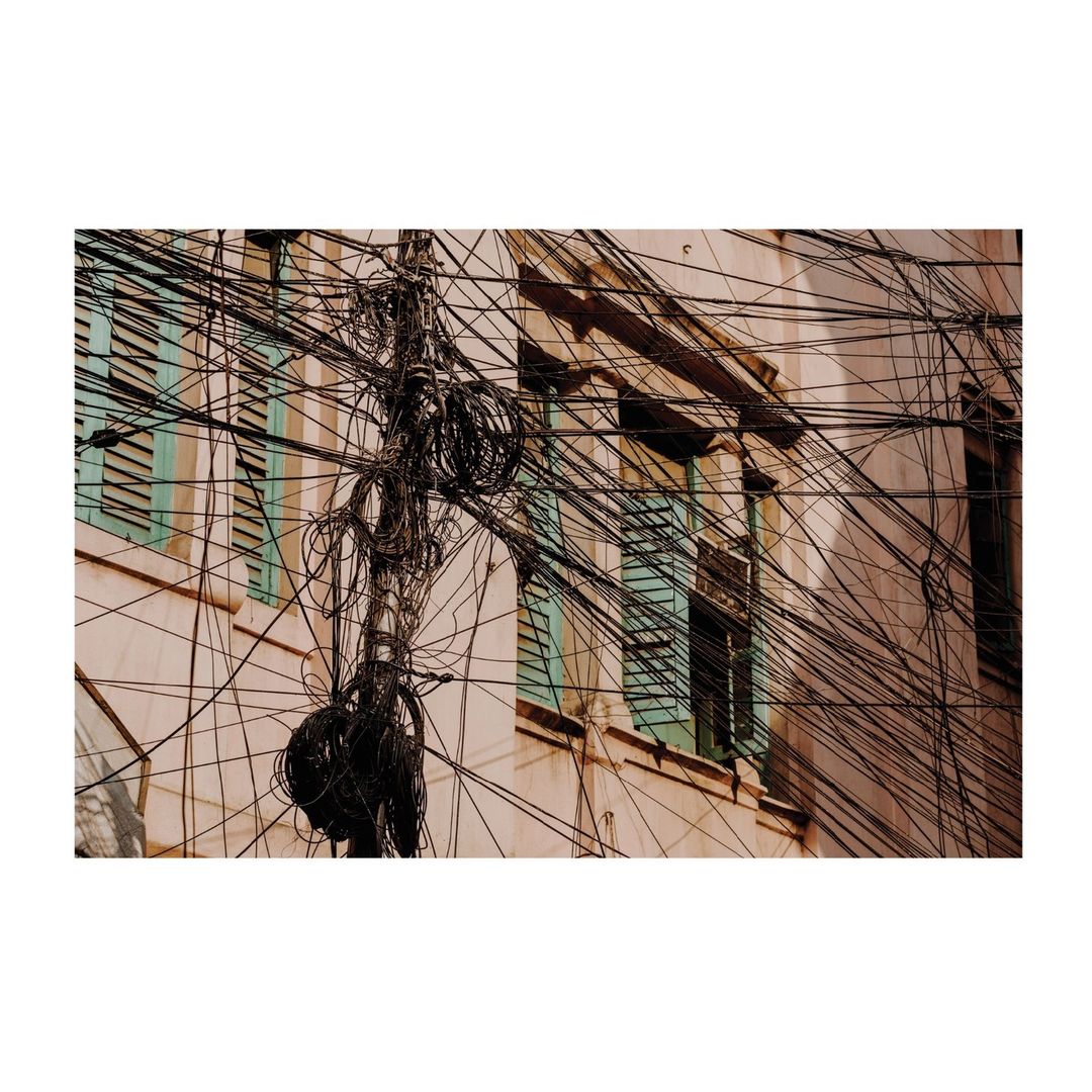 Tangled mess of overhead power lines obscures the view of a weathered building with windows, creating a chaotic urban scene.