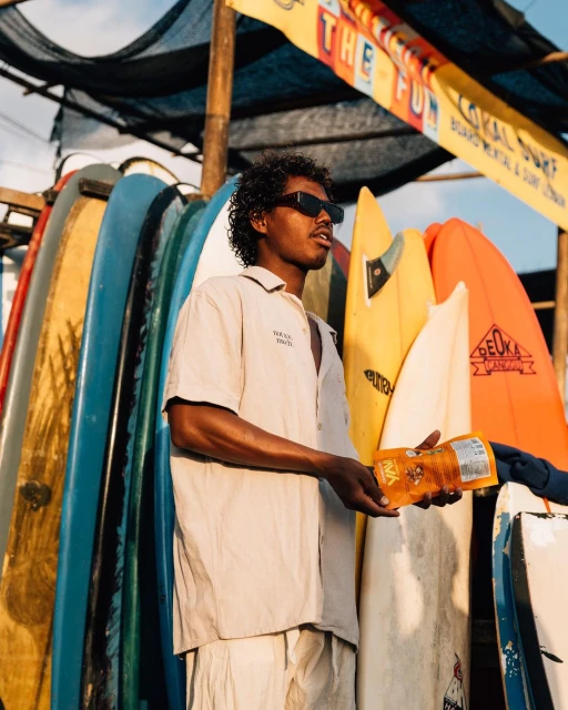 Young man with sunglasses stands near colorful surfboards, holding a snack package in outdoor lighting, creating a casual beach vibe.