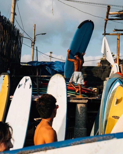 Man carries a blue surfboard over his head near a rack of other surfboards in an outdoor beach setting on a sunny day. 