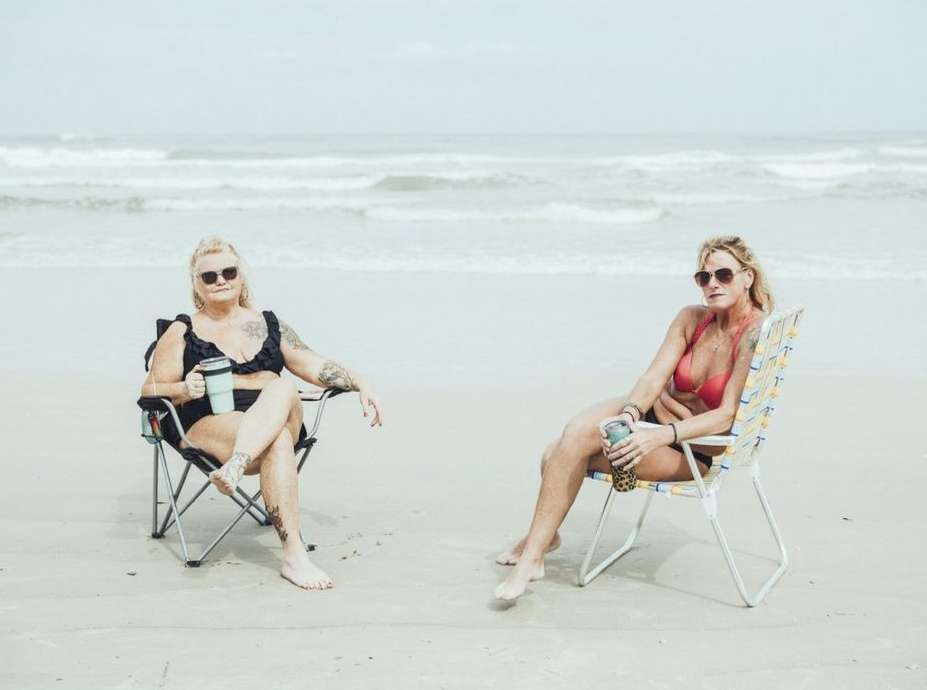 Two adult women in bikinis relax on beach chairs with drinks on a sandy beach near the ocean under an overcast sky.
